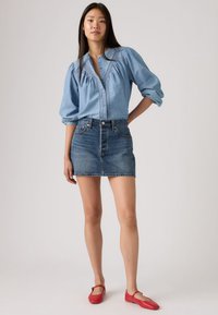 Ανοιχτό μπλε πουκάμισο από τζιν με φουσκωτά μανίκια και λεπτομερή ραφή, συνδυασμένο με σκούρο μπλε μίνι φούστα από τζιν και κόκκινα επίπεδα παπούτσια.