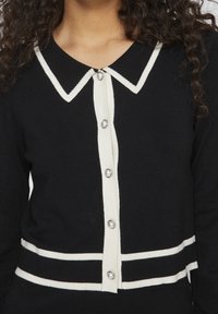 Svart kofta med vit krage och knappar. Anmärkningsvärda detaljer inkluderar en strukturerad stickning, horisontella vita ränder vid midjan, och pärlliknande knappar som accentuerar.