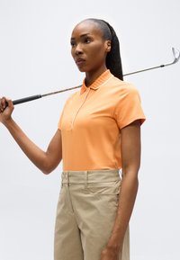 Femme tenant un club de golf reposant sur son épaule, portant un polo orange et un pantalon beige, sur un fond clair uni.