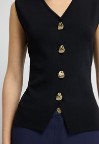 Granatowy top bez rękawów z dekoltem w serek i pięcioma nieregularnymi guzikami w kolorze złotym z przodu. Teksturowany materiał z dopasowanym krojem.