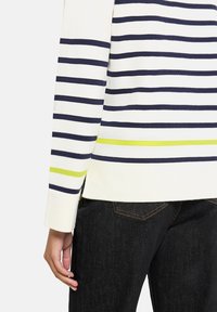 Długi rękaw w kremowym kolorze z granatowymi paskami, z wyrazistym zielonym akcentem przy krawędzi. Materiał wydaje się miękki, a krój dopasowany.