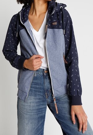 Persona che indossa una felpa con cappuccio con zip blu navy e grigia con motivi a ancora sulle maniche, maglietta bianca con scollo a V e jeans blu.