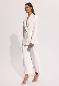 Kobieta ubrana w biały, dwurzędowy żakiet i szerokie spodnie, zestawione z sandałami na wysokim, cienkim obcasie, na jednolitym tle.