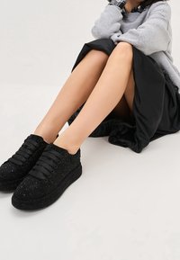 Czarne sneakersy o teksturowanym, nabijanym wzorze, z sznurowaniem i grubą, wzorzystą podeszwą. Noszone z czarną spódnicą i szarym topem.