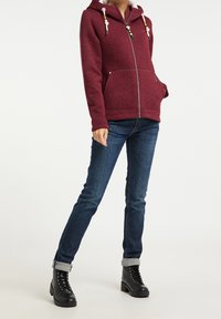 Bordowa polarowa bluza z kapturem, z zamkiem błyskawicznym z przodu i dekoracyjnymi sznurkami. Połączona z ciemnoniebieskimi dżinsami i czarnymi sznurowanymi butami.