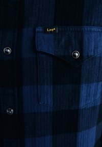 Cămașă din flanel cu model în carouri bleumarin și negru, cu o textură țesută, nasturi metalici cu clichet și o etichetă cu marcă pe buzunarul de la piept.