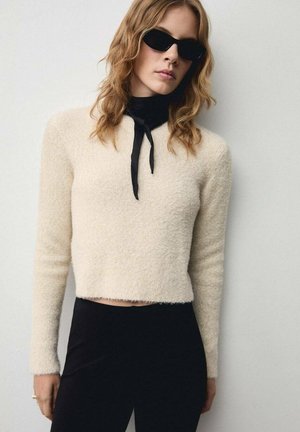 Gekürzter, cremefarbener Pullover aus flauschigem Material mit weicher Textur. Kombiniert mit einer schwarzen Krawatte und eleganten schwarzen Sonnenbrillen.