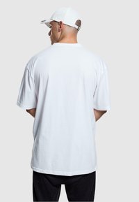 Weißes, übergroßes T-Shirt mit kurzen Ärmeln, das einen lässigen Schnitt und eine glatte Baumwoll Textur aufweist. Kombiniert mit schwarzen Hosen und einer weißen Kappe.