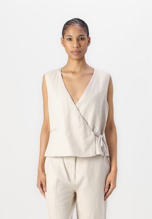 Top envolvente de lino beige con un profundo escote en V, diseño sin mangas y detalle de lazo lateral; textura suave y corte relajado combinado con pantalones a juego.