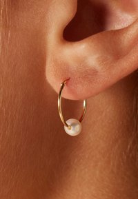 Gouden ringoorbel met een kleine witte parelaccent, die een gladde, gepolijste afwerking en een eenvoudig cirkelontwerp toont.