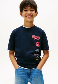 Camiseta de algodón azul marino con el logo "Tommy" en rojo, un emblema de "H" y varios parches en el lateral. Mangas cortas, cuello redondo.