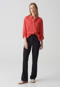 Camicia rossa a bottoni con due taschini sul petto, abbinata a pantaloni neri su misura e sneaker bianche con strisce rosse. Design semplice e pulito.