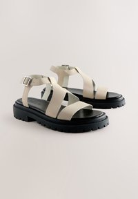 Beige læder sandaler med flere krydsede stropper, sort gummisål, justerbar ankelspænde og præget logo.