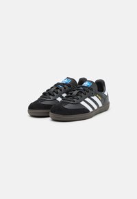 Baskets de sport noires avec rayures blanches, combinaison de daim et de cuir, semelle en caoutchouc et accents de logo bleus. Texte "SAMBA" en doré.