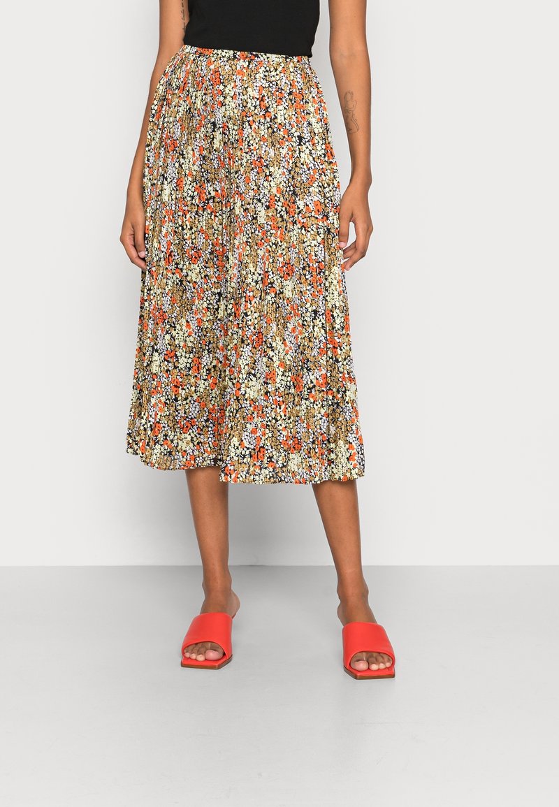 Femme portant une jupe midi plissée à motif floral orange, blanc et noir, un haut noir et des sandales slides à bout ouvert orange vif.