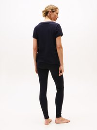 Kvinna stående barfota, vänd bort, iförd en marinblå kortärmad tröja och matchande marinblå leggings mot en ljus, enfärgad bakgrund.