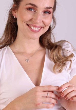 Zilveren hartketting met een heldere steen, gedragen met een witte V-hals blouse. Het model heeft lang golvend haar en laat een zilveren ring op haar hand zien.