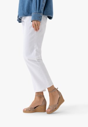 Onderbenen met witte cropped broeken en tan gekleurde sleehakken met enkelbandjes, gecombineerd met een spijkerjack mouw en een hand met ringen.