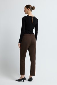 Vrouw in een zwarte top met lange mouwen en een rug-split, bruine slim-fit broek en zwarte schoenen met een lage hak, staand tegen een effen lichte muur.