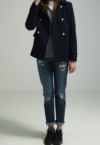 Marineblå dobbeltradet peacoat med guldknapper, grå sweater, slidte blå jeans og sorte sneakers. Opbrettede ærmer og afslappet pasform.