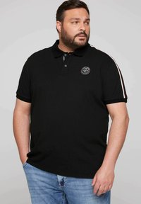 Czarna koszulka polo z kołnierzykiem i zapięciem na trzy guziki. Posiada okrągłą naszywkę z logo na piersi oraz kontrastowe paski na rękawach.