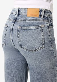 Hellblaue, hochtaillierte Jeans mit Gesäßtaschen und einem beigen Leder-Brandpatch mit der Aufschrift „NOISY MAY“, getragen zu einem weißen Top.