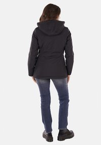 Giacca leggera nera con cappuccio, caratterizzata da un design a vita svasata, texture liscia e maniche lunghe, abbinata a jeans blu e scarpe nere.