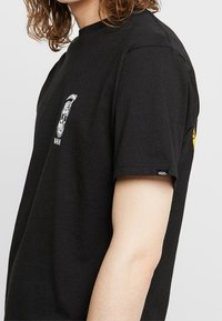 Musta puuvillainen t-paita, jossa valkoinen graafinen kuvio rinnassa ja keltainen korostus hihassa. Lyhyet hihat ja pyöreä kaula-aukko.