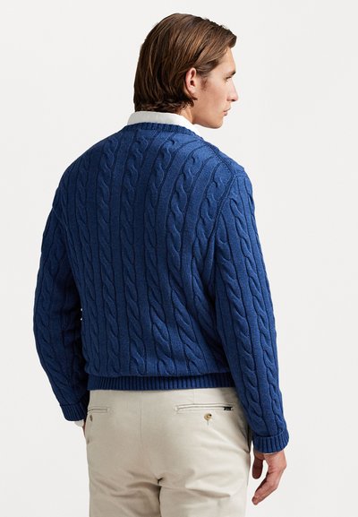 Μπλε πουλόβερ με πλέξη καλωδίου και υφασμάτινο σχέδιο, συνδυασμένο με ανοιχτό μπεζ παντελόνι που διαθέτει λεπτομέρειες στην πίσω τσέπη.