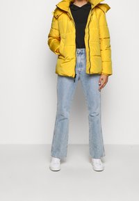 Gele gewatteerde jas met rits aan de voorkant, capuchon en zwarte accenten, gedragen over een zwart shirt, gecombineerd met lichtblauwe wijde spijkerbroeken en witte sneakers.