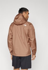 Vandtæt brun jakke med hætte, der har elastiske manchetter og The North Face-logoet på den øverste ryg. Bæres med sorte shorts.