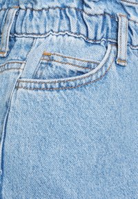 Ljust blå denimtyg med ett rynkat midjeband, som har en framficka och kontrasterande orange sömdetaljer.