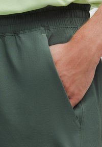 Tumši zaļi sporta šorti ar elastīgu jostasvietu, ar sānu kabatām un gludu tekstūru, daļēji parādīti ar ielikto roku.