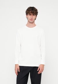 Fehér hosszú ujjú ing bordázott textúrával és kör alakú nyakkivágással. Sötét nadrággal párosítva, minimális márkajelzéssel a mellrészen.