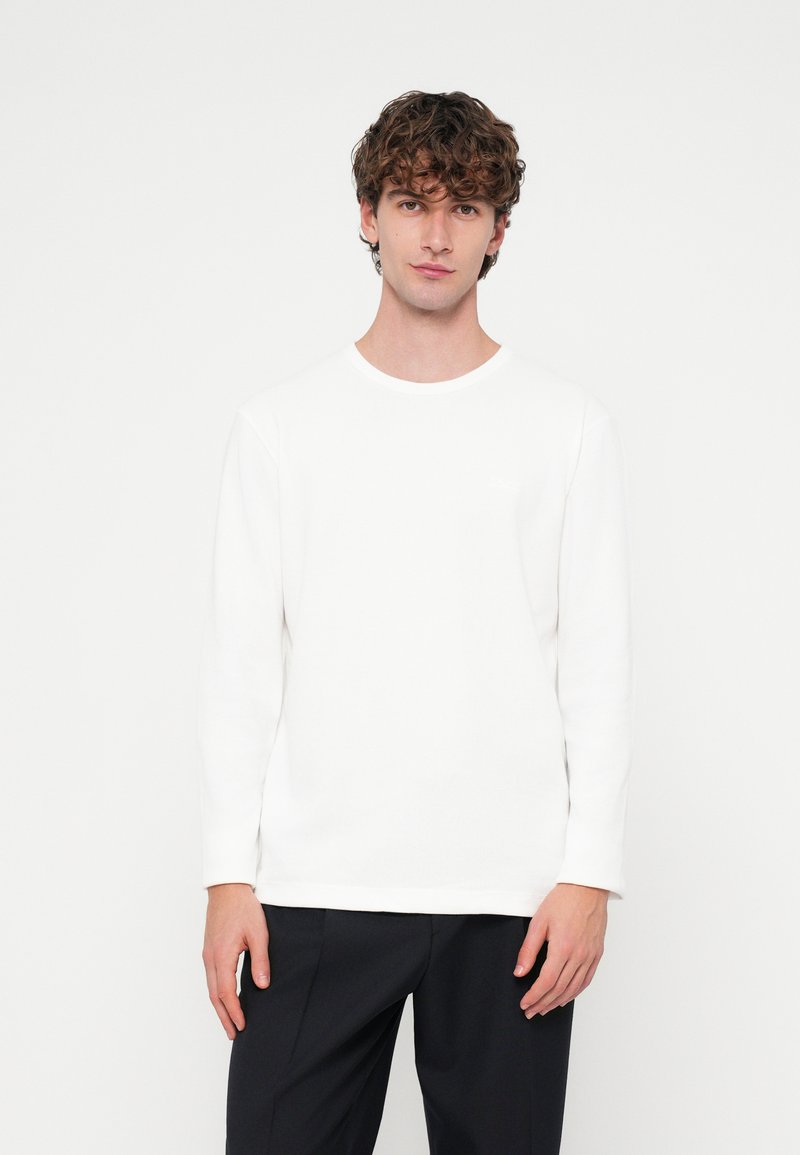 Fehér hosszú ujjú ing bordázott textúrával és kör alakú nyakkivágással. Sötét nadrággal párosítva, minimális márkajelzéssel a mellrészen.