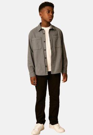 Grå skjorte med knapper og to brystlommer, båret over en hvid T-shirt, parret med sorte bukser og hvide sneakers. Ensfarvet baggrund.