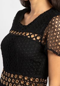 Robe en dentelle noire présentant un motif texturé, des manches courtes et une bande horizontale d'œillets dorés à la taille pour un détail remarquable.