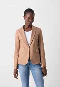 Béžový přiléhavý blazer s jedním knoflíkem, dvěma kapsami a hladkou texturou, nošený přes bílé tričko a modré džínsy.