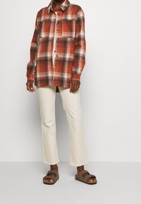 Geruit overhemd in rood, wit en zwart flanel, met knopen aan de voorkant, gedragen met licht crèmekleurige broek en bruine sandalen. Casual stijl.