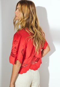 Rote bestickte Bluse mit gewellten Kanten und Ausschnittmustern. Das Model trägt sie über hellbeigen, strukturierten Hosen. Die Haare fallen herab.