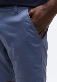 Σκούρο δέρμα χέρι που ακουμπά στην εμπρόσθια τσέπη μπλε casual σορτς, με φόντο απλό και ανοιχτόχρωμο.