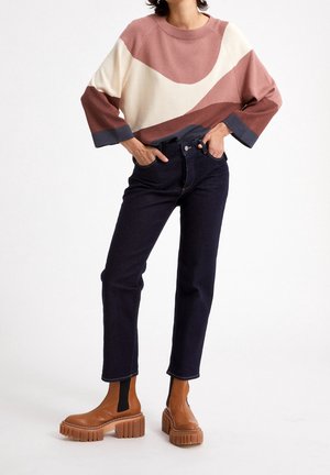 Person iført color-blocked sweater i pink, creme og brun, mørke jeans og chunky brune plateau-støvler, stående med hænderne i lommerne.