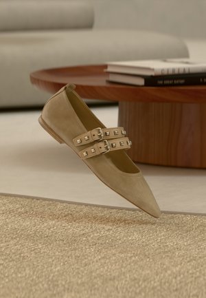 Chaussure plate en daim beige avec un bout pointu, dotée de deux brides réglables ornées de clous métalliques. Légèrement surélevée au-dessus d'une surface texturée.