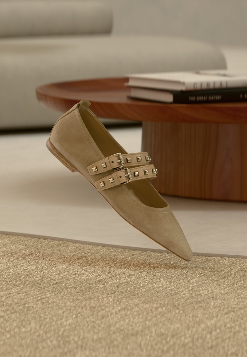 Μπεζ φλατ παπούτσι από σουέτ με μυτερή μύτη, που διαθέτει δύο ρυθμιζόμενες λουράκια διακοσμημένα με μεταλλικά καρφάκια. Ελαφρώς ανυψωμένο πάνω σε υφή επιφάνειας.
