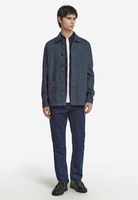 Mörkblå lättviktsjacka med krage, knappstängning och flera fickor, som bärs över en vit skjorta och blå jeans.