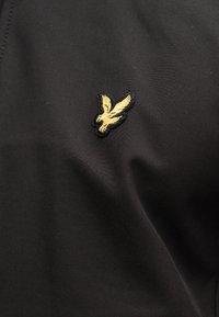 Μαύρο ύφασμα με μικρό χρυσό κέντημα που απεικονίζει πουλί σε μέση πτήση, με λεπτομερείς φτερούγες και ράμφος που κοιτάζει προς τα κάτω.