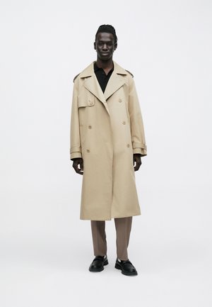 Nuori mies, jolla on tumma iho ja punotut hiukset, pukeutuneena beigeen trenssitakkiin, mustaan paitaan, ruskeisiin housuihin ja mustiin kenkiin, seisoo valkoisella taustalla.