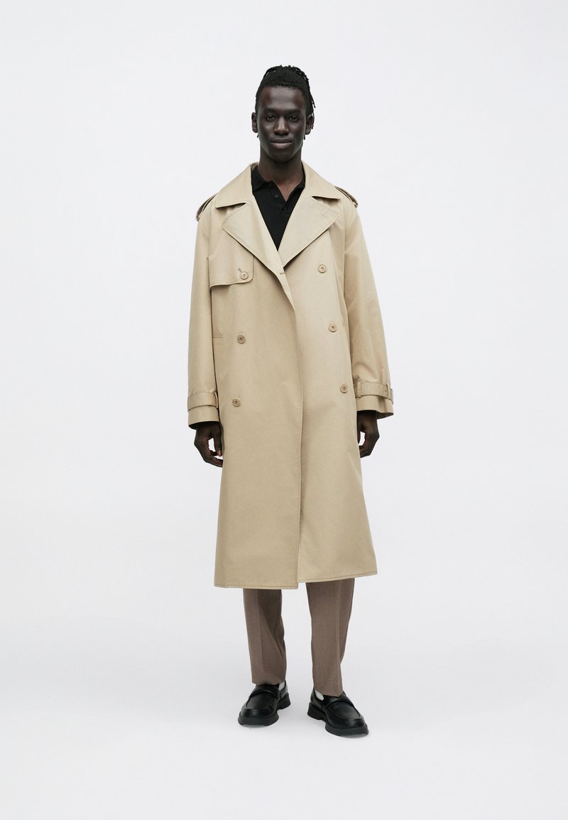 Jeune homme à la peau foncée et aux cheveux tressés portant un trench-coat beige, une chemise noire, un pantalon marron et des chaussures noires, debout sur un fond blanc.