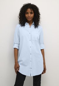 Vaaleansininen nappipaita, jossa on terävä kaulus, yksittäinen rintatasku ja käärityt hihat. Paidassa on kaareva helma ja pehmeä kangaspinta.