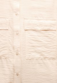 Camisa beige clara con textura arrugada, dos bolsillos en el pecho y una tapeta con botones de perla. Tejido suave y ligero.